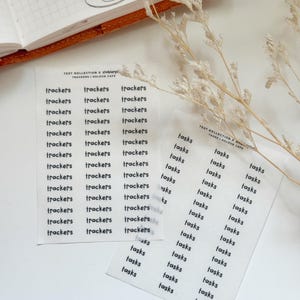 May include: Two sheets of white stickers with black text. One sheet says "trackers" repeatedly, and the other says "tasks." The stickers are from the "TEXT KOLLECTION x stebierpi TRACKERS | KOLOUR CAFE" collection.