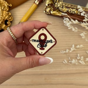 May include: A diamond-shaped patch with a cream background and a maroon border. The patch features the text "Traveller's Club" and a circular design. The patch is being held by a person.