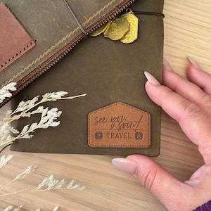 Peut inclure: Un journal de voyage en cuir marron foncé avec une fermeture éclair et un patch marron avec l'inscription "see you soon / TRAVEL". Le journal a des breloques dorées et un accent en cuir marron.