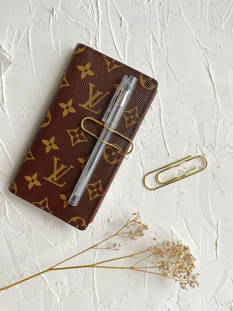 Puede incluir: Una libreta de cuero marr&oacute;n con un patr&oacute;n de monograma dorado, un bol&iacute;grafo transparente y dos clips de papel dorados sobre una superficie blanca texturizada. Una ramita de flores secas se encuentra en la esquina inferior derecha.