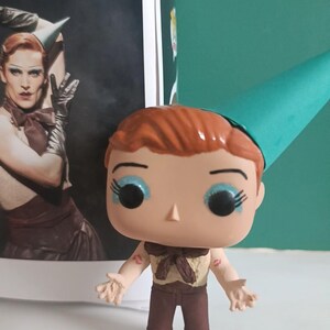 May include: A Funko Pop vinyl figure of a character with red hair, blue eye shadow, and a teal party hat. The figure is wearing a brown outfit and has lipstick marks on its arms. A photo of a person in drag is in the background.