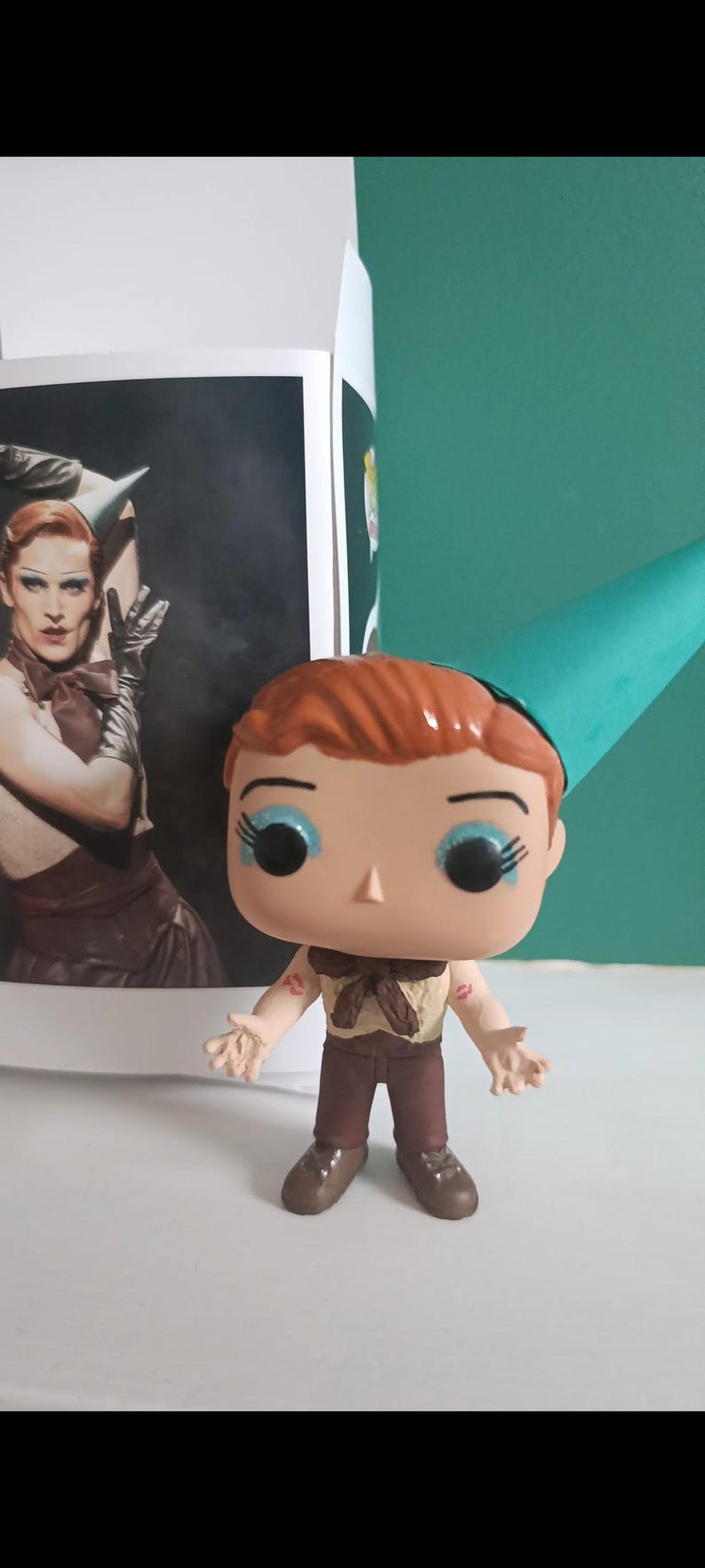 May include: A Funko Pop vinyl figure of a character with red hair, blue eye shadow, and a teal party hat. The figure is wearing a brown outfit and has lipstick marks on its arms. A photo of a person in drag is in the background.