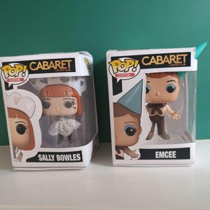 May include: Two Pop! Custom vinyl figures from the musical Cabaret, Sally Bowles and Emcee, in their original boxes. Sally Bowles is wearing a white dress and hat. Emcee is wearing a brown outfit and a blue hat. The boxes have the words "Cabaret" and "Kit Kat Club".