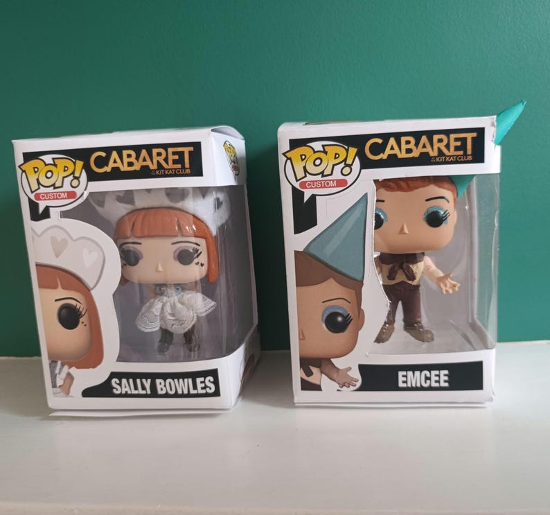 May include: Two Pop! Custom vinyl figures from the musical Cabaret, Sally Bowles and Emcee, in their original boxes. Sally Bowles is wearing a white dress and hat. Emcee is wearing a brown outfit and a blue hat. The boxes have the words "Cabaret" and "Kit Kat Club".