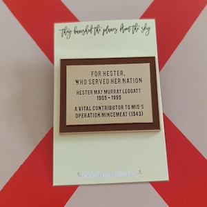 May include: A rectangular pin with a brown border and a gold-coloured centre, featuring text honouring Hester May Murray Leggatt. The pin is on a cream-coloured card with the words "they banished the planes from the sky" at the top.