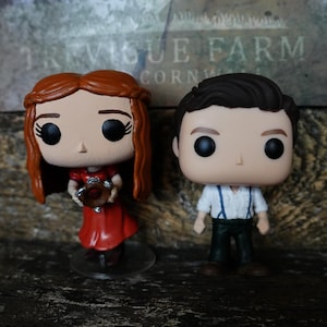 Könnte beinhalten: Zwei Funko Pop Figuren. Eine Figur ist eine Frau in einem roten Kleid, die ein braunes Objekt hält. Die andere Figur ist ein Mann in einem weißen Hemd, dunkler Hose und blauen Hosenträgern. Im Hintergrund steht "TREVIGUE FARM CORNWALL".