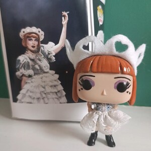 May include: A Funko Pop figure of a character with red hair, wearing a white dress and a white crown. The figure is next to a box with a picture of the character. The figure has black boots and large eyes.