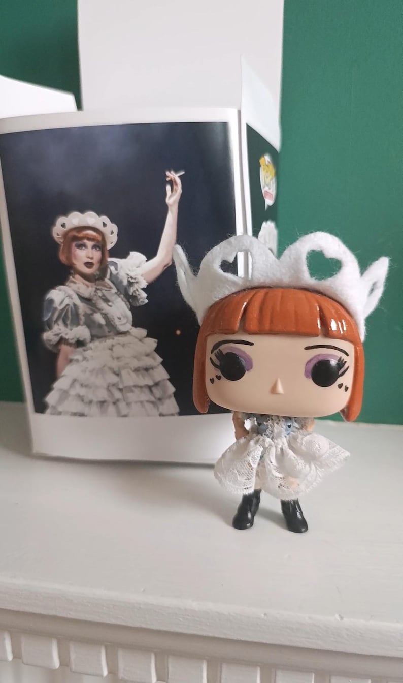 May include: A Funko Pop figure of a character with red hair, wearing a white dress and a white crown. The figure is next to a box with a picture of the character. The figure has black boots and large eyes.