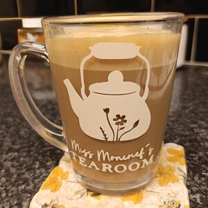 May include: A glass mug containing a light brown drink, decorated with a white teapot motif and the words "Miss Moniref's Tearoom." The mug sits on a floral coaster with yellow flowers and a bee.