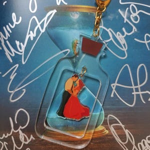 May include: Clear acrylic keychain in the shape of a bottle, featuring a couple embracing. The keychain has a gold-colored clasp and chain. The background is a blue image with white signatures and the text "THE CURIOUS CASE OF BENJAMIN BUTTON".
