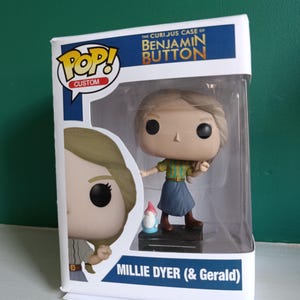 Könnte beinhalten: Eine benutzerdefinierte Pop! Vinyl-Figur von Millie Dyer aus "Der seltsame Fall des Benjamin Button". Die Figur trägt ein grün-gelb gestreiftes Hemd, einen blauen Rock und braune Stiefel. Eine kleine Zwergfigur ist enthalten. Die Box zeigt das Pop!-Logo und den Namen der Figur.