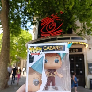 May include: A Pop! Custom figure of Emcee from Cabaret in a clear plastic display case. The figure has red hair, blue eye makeup, and wears a tan shirt, brown pants, and a blue hat. The box reads "CABARET & KIT KAT CLUB" and "EMCEE".