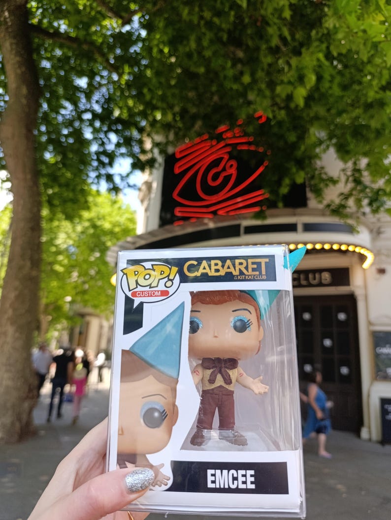 May include: A Pop! Custom figure of Emcee from Cabaret in a clear plastic display case. The figure has red hair, blue eye makeup, and wears a tan shirt, brown pants, and a blue hat. The box reads "CABARET & KIT KAT CLUB" and "EMCEE".