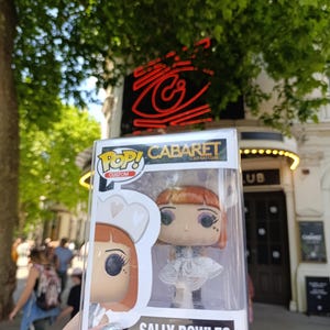 May include: A Funko Pop! figure of Sally Bowles from Cabaret, in a clear plastic display case. The figure has red hair, a white dress, and black boots. The box reads "CABARET" and "SALLY BOWLES". The background shows a theater.
