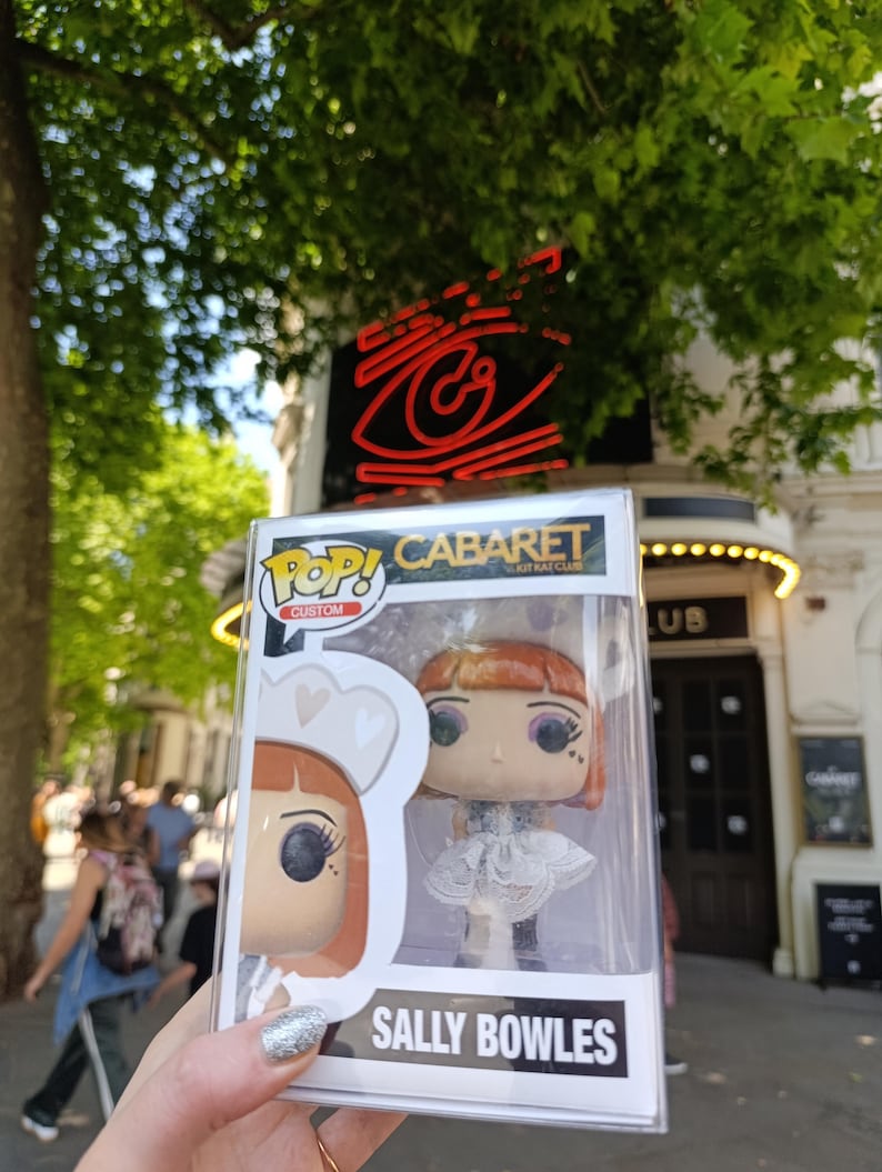 May include: A Funko Pop! figure of Sally Bowles from Cabaret, in a clear plastic display case. The figure has red hair, a white dress, and black boots. The box reads "CABARET" and "SALLY BOWLES". The background shows a theater.