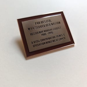 May include: A rectangular bronze plaque with a dark brown border. The plaque features engraved text honouring Hester, including her name, dates (1905-1995), and her contribution to Operation Mincemeat.