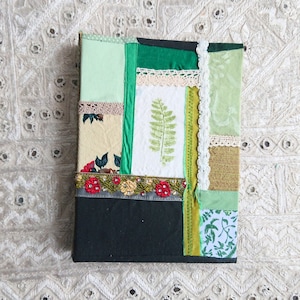 Può includere: Un diario fatto a mano con una copertina in patchwork verde, marrone e bianco. La copertina è decorata con pizzo, nastro e un motivo a felce.