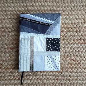Puede incluir: Una cubierta de cuaderno de patchwork gris y blanco con detalles de encaje y cinta. La cubierta está hecha de diferentes telas y texturas, incluyendo un fieltro gris, algodón blanco y tela estampada en blanco y negro.