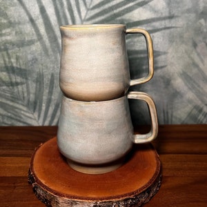 Könnte beinhalten: Zwei übereinander gestapelte Keramikbecher mit hellgrauer Glasur. Die Becher haben einen abgerundeten Körper und einen Henkel. Die Becher stehen auf einem Holzuntersetzer.