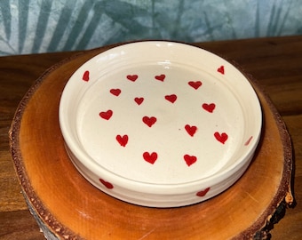 May include: A circular, cream-coloured ceramic dish with a rim, adorned with small, evenly spaced red hearts. The dish rests on a natural wood slice, giving a rustic feel. The background features a textured, teal-coloured wall.