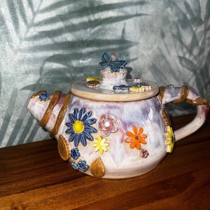 Könnte beinhalten: Eine Keramik-Teekanne mit lila Glasur und floralen Akzenten. Die Teekanne hat einen braunen Henkel und einen Deckel mit einem Schmetterlingsdesign. Die Teekanne ist mit Blumen, Blättern und einem Schmetterling verziert.