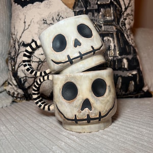 Könnte beinhalten: Zwei gestapelte Keramikbecher mit cremefarbener Glasur und handgemaltem Totenkopf-Design. Jeder Becher hat tiefe schwarze Augenhöhlen, eine dreieckige Nase und einen genähten Mund. Die Henkel sind mit einem schwarz-weißen Streifenmuster versehen.