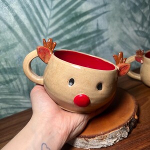 May include: A handmade ceramic reindeer mug with a tan exterior, red interior, and a red nose. The mug features small antlers and ears. The mug is sitting on a wooden coaster. The background is a green and blue leaf pattern.