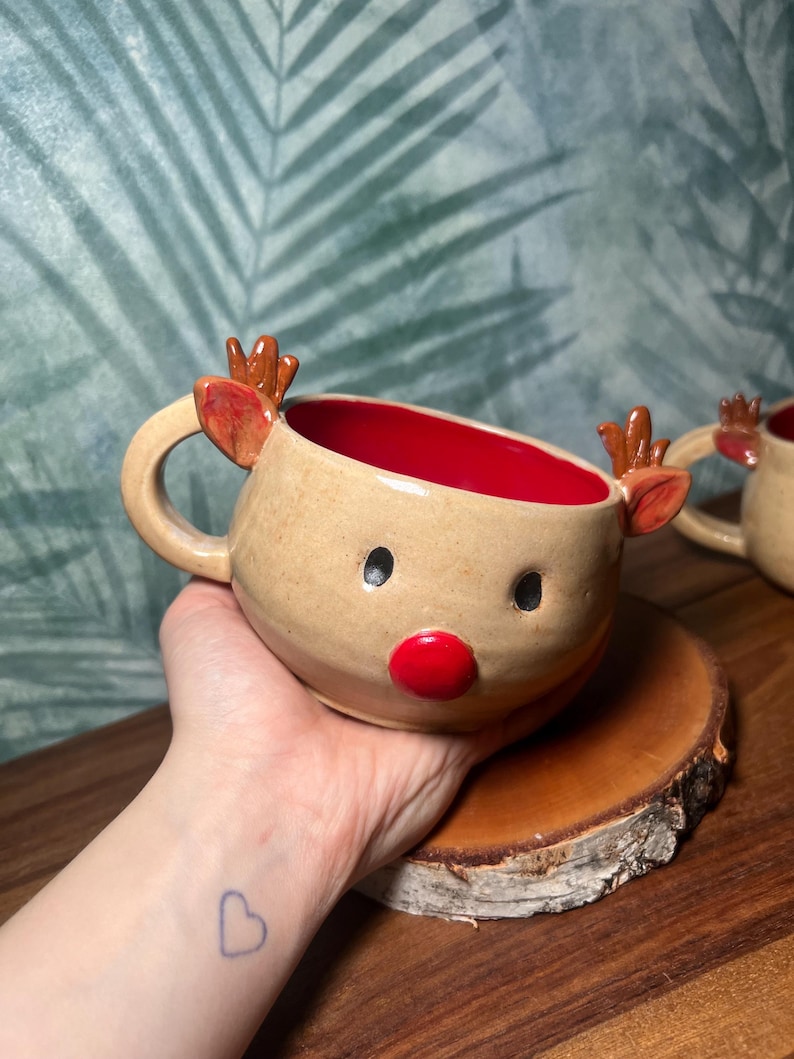 May include: A handmade ceramic reindeer mug with a tan exterior, red interior, and a red nose. The mug features small antlers and ears. The mug is sitting on a wooden coaster. The background is a green and blue leaf pattern.