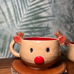 May include: A ceramic reindeer mug with a tan exterior, red interior, and a red nose. The mug features brown antlers and black eye details. It sits on a wooden coaster, perfect for holiday beverages.