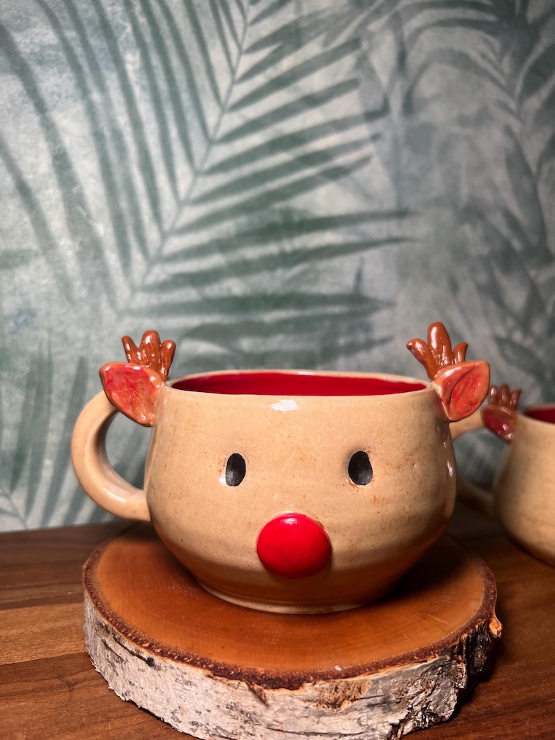 May include: A ceramic reindeer mug with a tan exterior, red interior, and a red nose. The mug features brown antlers and black eye details. It sits on a wooden coaster, perfect for holiday beverages.