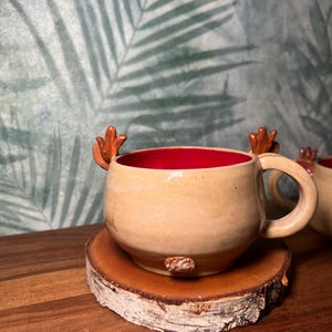 May include: A handmade ceramic mug with a tan exterior and a red interior. The mug features antler-shaped handles and sits on a wooden coaster. The mug is designed for hot beverages.