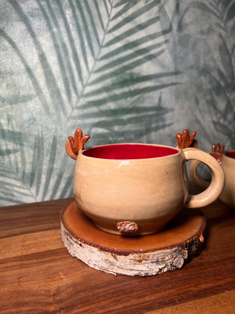 May include: A handmade ceramic mug with a tan exterior and a red interior. The mug features antler-shaped handles and sits on a wooden coaster. The mug is designed for hot beverages.
