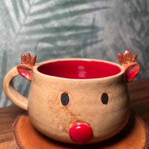 May include: A handmade ceramic mug shaped like a reindeer. The mug has a tan exterior with a red interior. It features black eyes, a red nose, and small antlers. The mug is sitting on a wooden coaster.