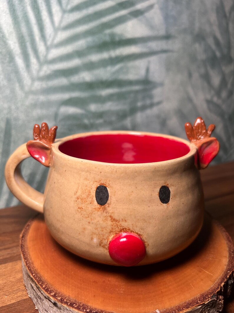 May include: A handmade ceramic mug shaped like a reindeer. The mug has a tan exterior with a red interior. It features black eyes, a red nose, and small antlers. The mug is sitting on a wooden coaster.