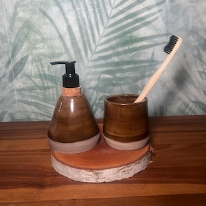 Puede incluir: Un dispensador de jabón de cerámica marrón con tapa de corcho y un portacepillos de dientes de cerámica marrón a juego con un cepillo de dientes negro. Los artículos están sobre una superficie de madera.
