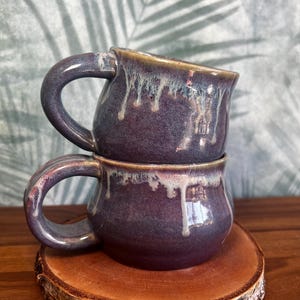 Könnte beinhalten: Zwei handgefertigte Keramikbecher, die übereinander gestapelt sind. Die Becher sind tiefviolett mit einem tropfenden Glanzeffekt in Weiß und Beige. Jeder Becher hat einen abgerundeten Henkel. Die Becher stehen auf einem runden Holzuntersetzer.