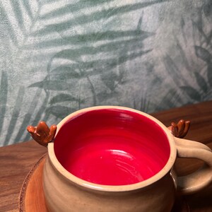 May include: A beige ceramic mug with a red interior, featuring small brown antler-shaped handles. The mug sits on a wooden coaster, with a blurred background of green and white foliage.
