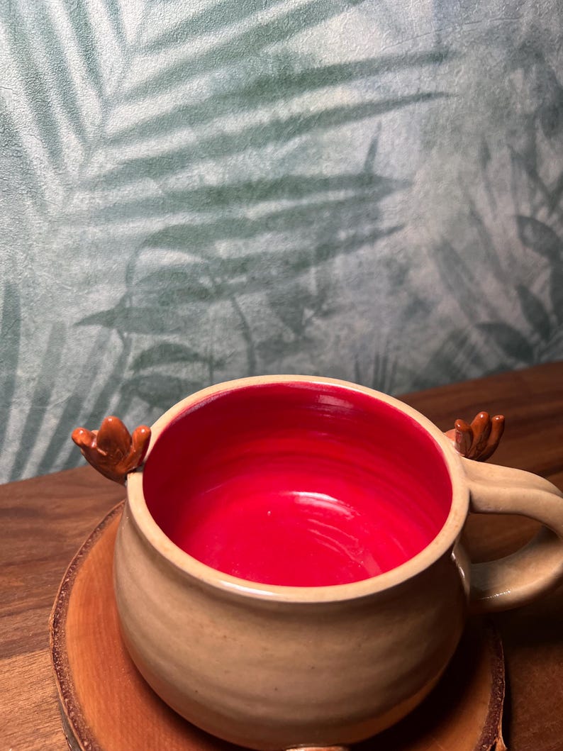 May include: A beige ceramic mug with a red interior, featuring small brown antler-shaped handles. The mug sits on a wooden coaster, with a blurred background of green and white foliage.