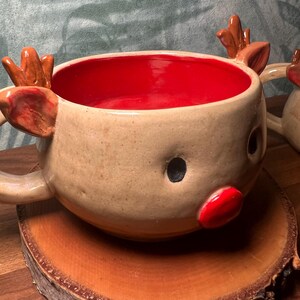 May include: A handmade ceramic reindeer mug with a tan exterior, red interior, and brown antlers. The mug features a red nose and black eyes, perfect for holiday beverages. The mug is sitting on a wooden slice.
