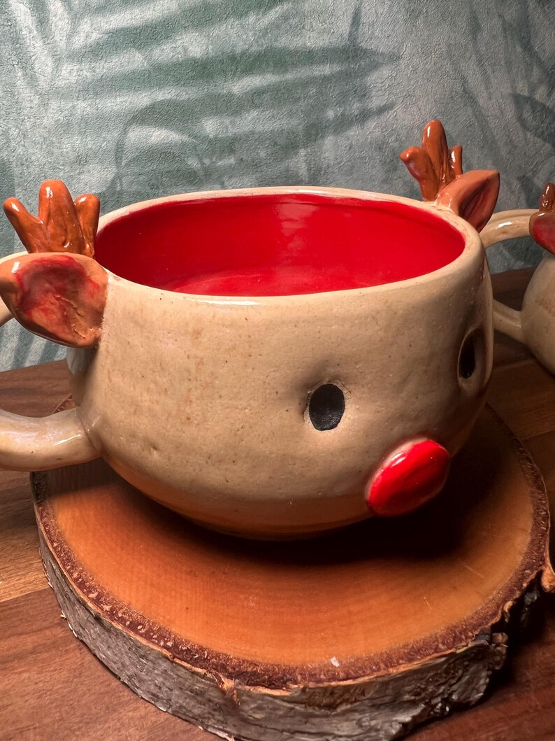 May include: A handmade ceramic reindeer mug with a tan exterior, red interior, and brown antlers. The mug features a red nose and black eyes, perfect for holiday beverages. The mug is sitting on a wooden slice.
