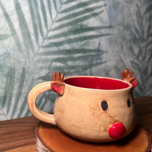 May include: A ceramic mug shaped like a reindeer, with a tan exterior and a red interior. The mug features a red nose, black eyes, and small brown antlers. It sits on a wooden coaster.