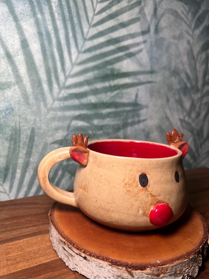 May include: A ceramic mug shaped like a reindeer, with a tan exterior and a red interior. The mug features a red nose, black eyes, and small brown antlers. It sits on a wooden coaster.
