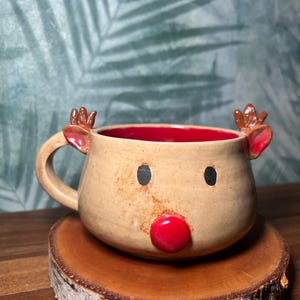 May include: A handmade ceramic mug shaped like a reindeer, with a tan exterior, red interior, and a red nose. The mug has black eyes and small antlers. It sits on a wooden slice.