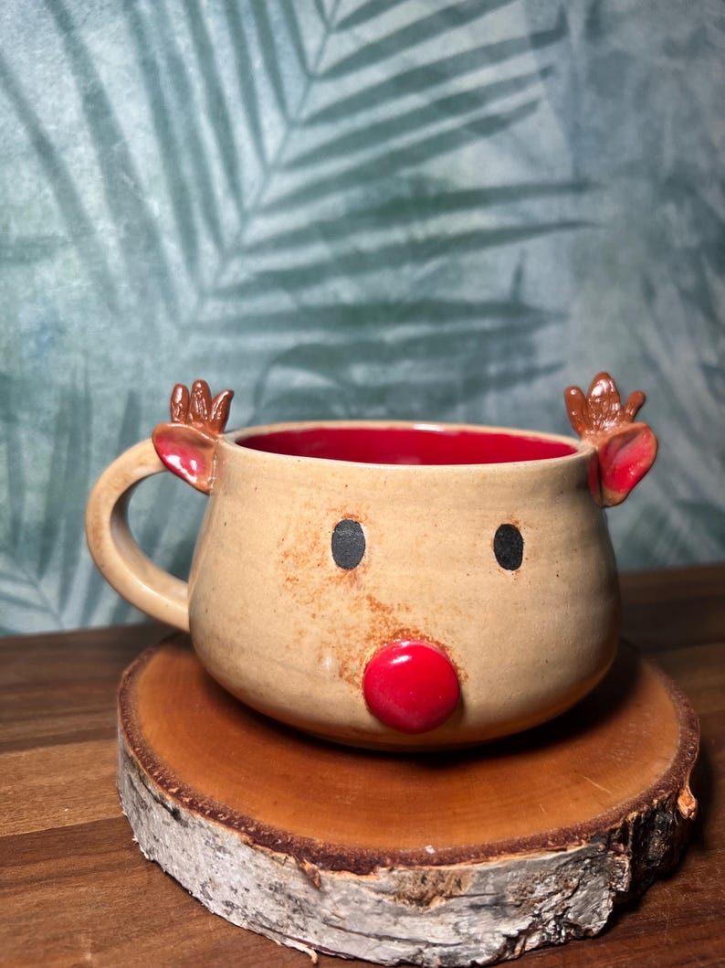 May include: A handmade ceramic mug shaped like a reindeer, with a tan exterior, red interior, and a red nose. The mug has black eyes and small antlers. It sits on a wooden slice.