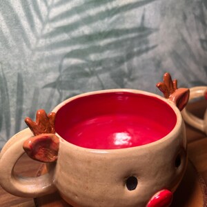 May include: A handmade ceramic reindeer mug with a tan exterior and a bright red interior. The mug features a reindeer face with black eyes, a red nose, and small antlers. The mug is designed for hot beverages.