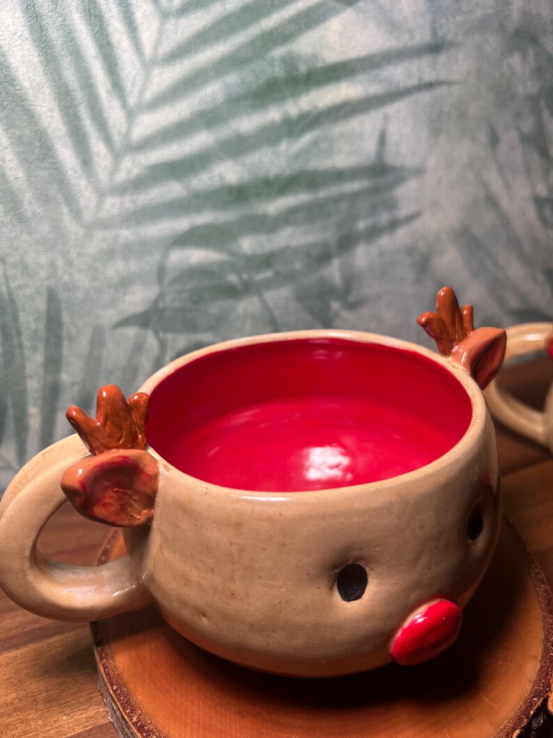 May include: A handmade ceramic reindeer mug with a tan exterior and a bright red interior. The mug features a reindeer face with black eyes, a red nose, and small antlers. The mug is designed for hot beverages.
