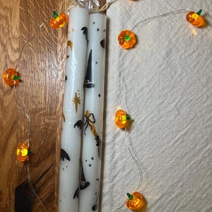 May include: Two white Halloween candles decorated with black witch hats, bats, and stars. A string of orange pumpkin lights is wrapped around the candles. The candles are packaged in clear cellophane.