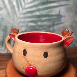 May include: A handmade ceramic mug in the shape of a reindeer. The mug has a tan exterior with a red interior. It features black dot eyes, small brown antlers, and a red nose. The mug is sitting on a wooden coaster.