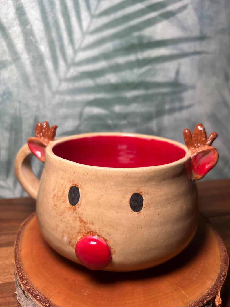 May include: A handmade ceramic mug in the shape of a reindeer. The mug has a tan exterior with a red interior. It features black dot eyes, small brown antlers, and a red nose. The mug is sitting on a wooden coaster.