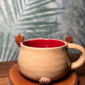 May include: A tan ceramic mug with a red interior, featuring small brown antler-like handles and a textured brown nose. The mug is placed on a round wooden coaster, perfect for coffee or tea.